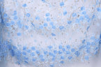a close-up view of a delicate, blue lace fabric adorned with a floral pattern. The lace is intricately embroidered with numerous blue roses and leaves, creating a visually appealing and delicate design. The fabric appears to be a light blue color, which complements the blue roses and leaves, enhancing the overall aesthetic of the piece.