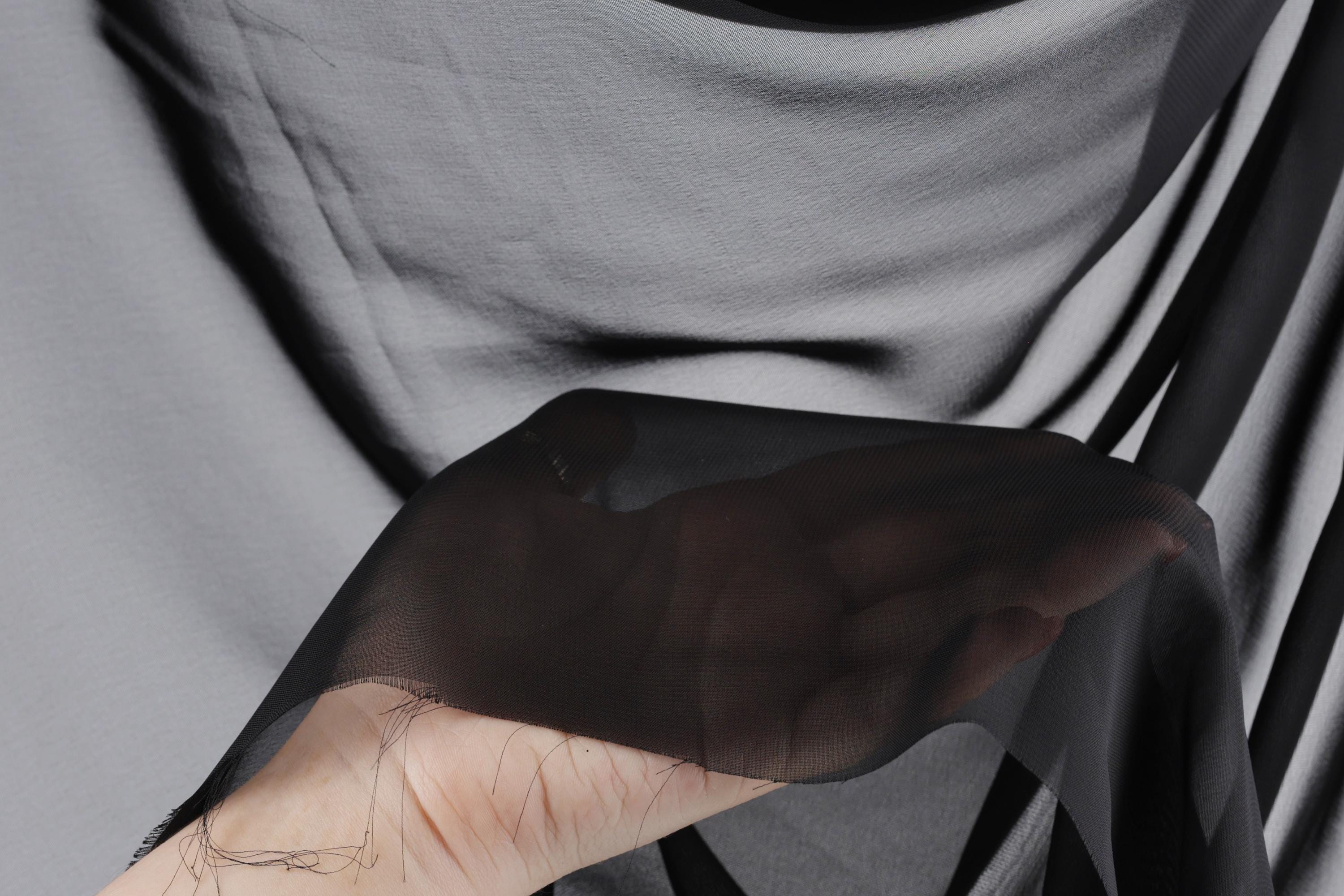 A hand is holding a piece of black fabric, which appears to be a sheer material. The background is a dark gray color, creating a contrast with the hand and fabric.