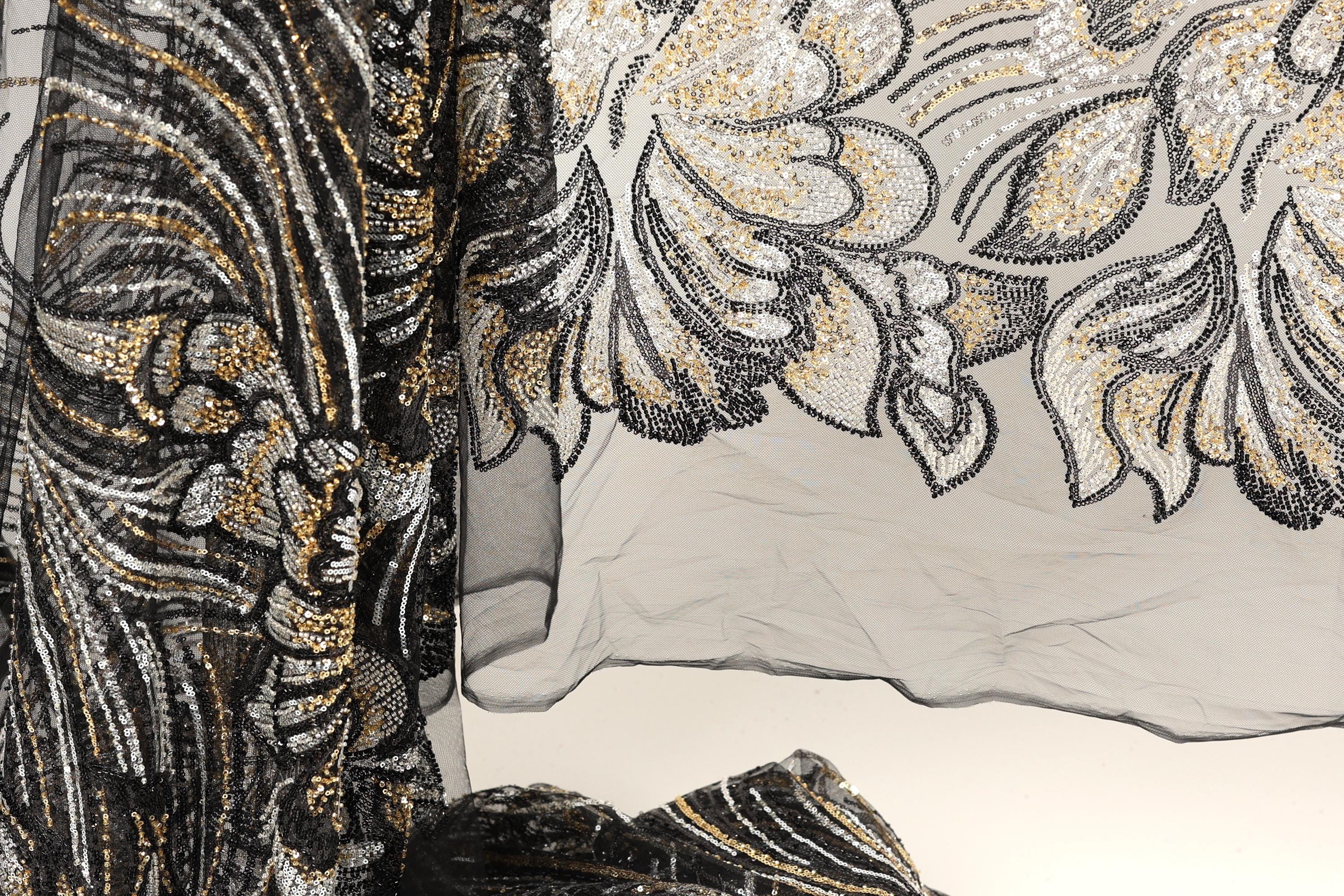 a close-up view of a black and gold sequined fabric with a floral pattern. The fabric appears to be a sheer material, with a black and gold color scheme. The image does not contain any text or other discernible objects or actions.