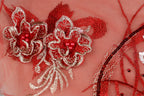 a close-up view of a delicate, intricately embroidered floral design on a pink background. The design is composed of two large red flowers with white and silver accents, including small red beads and a metallic ribbon. The background is a soft pink color, which complements the vibrant red of the flowers and beads. The image does not contain any text or other discernible objects, and the focus is solely on the detailed embroidery work.