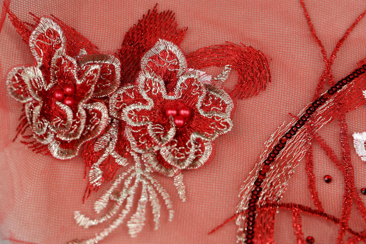 a close-up view of a delicate, intricately embroidered floral design on a pink background. The design is composed of two large red flowers with white and silver accents, including small red beads and a metallic ribbon. The background is a soft pink color, which complements the vibrant red of the flowers and beads. The image does not contain any text or other discernible objects, and the focus is solely on the detailed embroidery work.