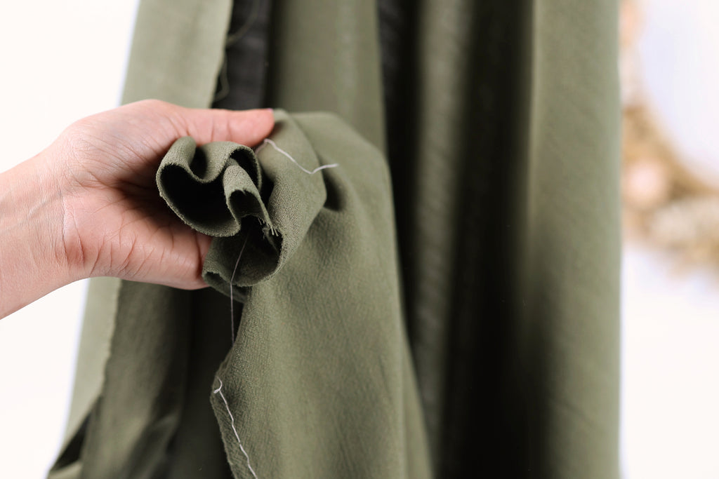 A hand holding a piece of green fabric with a white thread visible, against a blurred background.