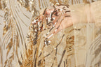 A hand is holding a piece of fabric with gold and black sequin patterns, against a background of gold and black sequin designs.