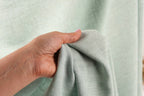 A hand is holding a piece of light green fabric, possibly a curtain or textile, in front of a blurred background.