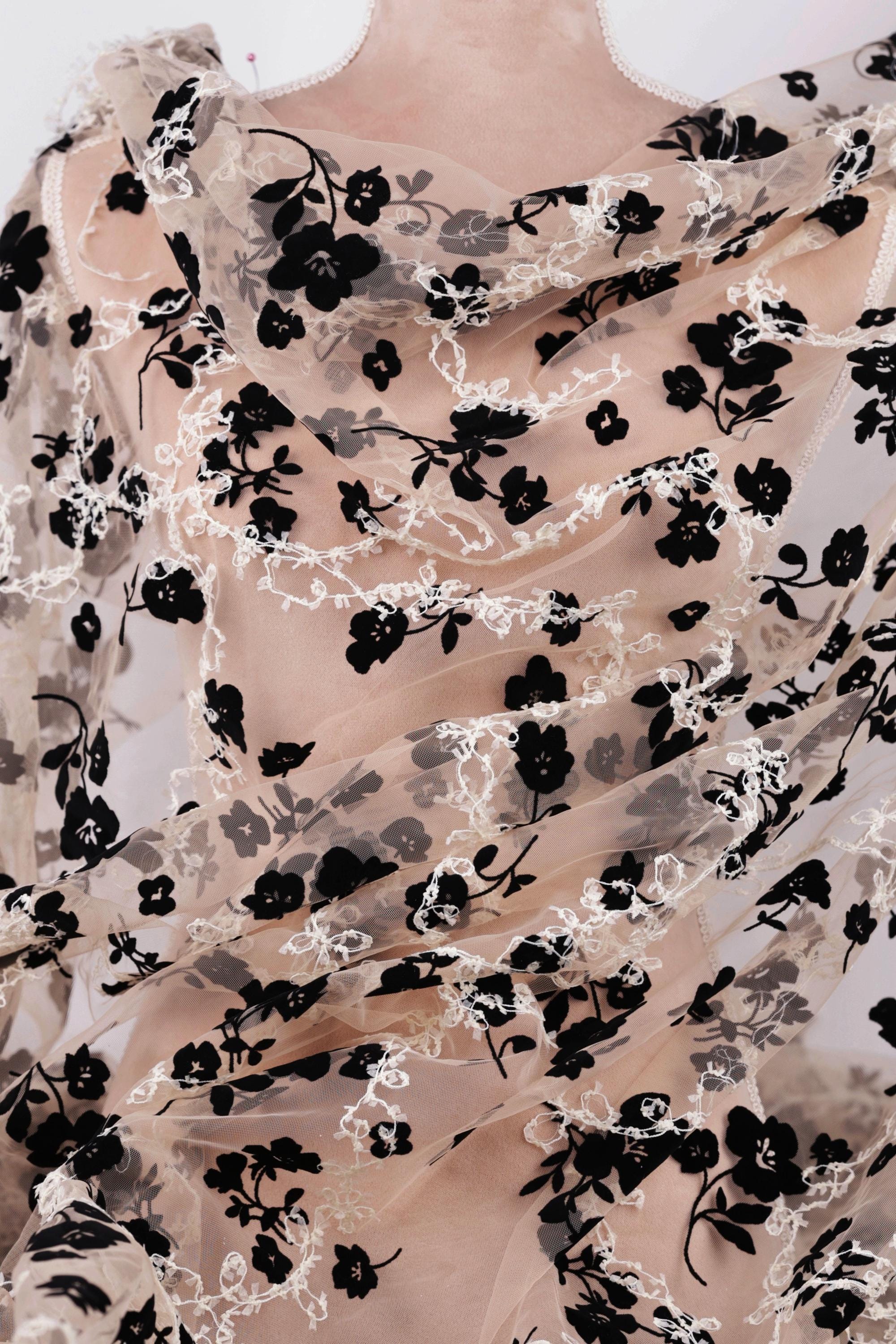 a close-up view of a sheer fabric with a black floral pattern. The fabric appears to be made of sheer material, giving it a delicate and airy appearance. The black flowers are arranged in a way that covers the entire fabric, creating a visually striking contrast against the lighter background.
