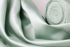 A close-up view of a soft, mint green satin fabric, with a rolled-up piece of the fabric visible in the background.