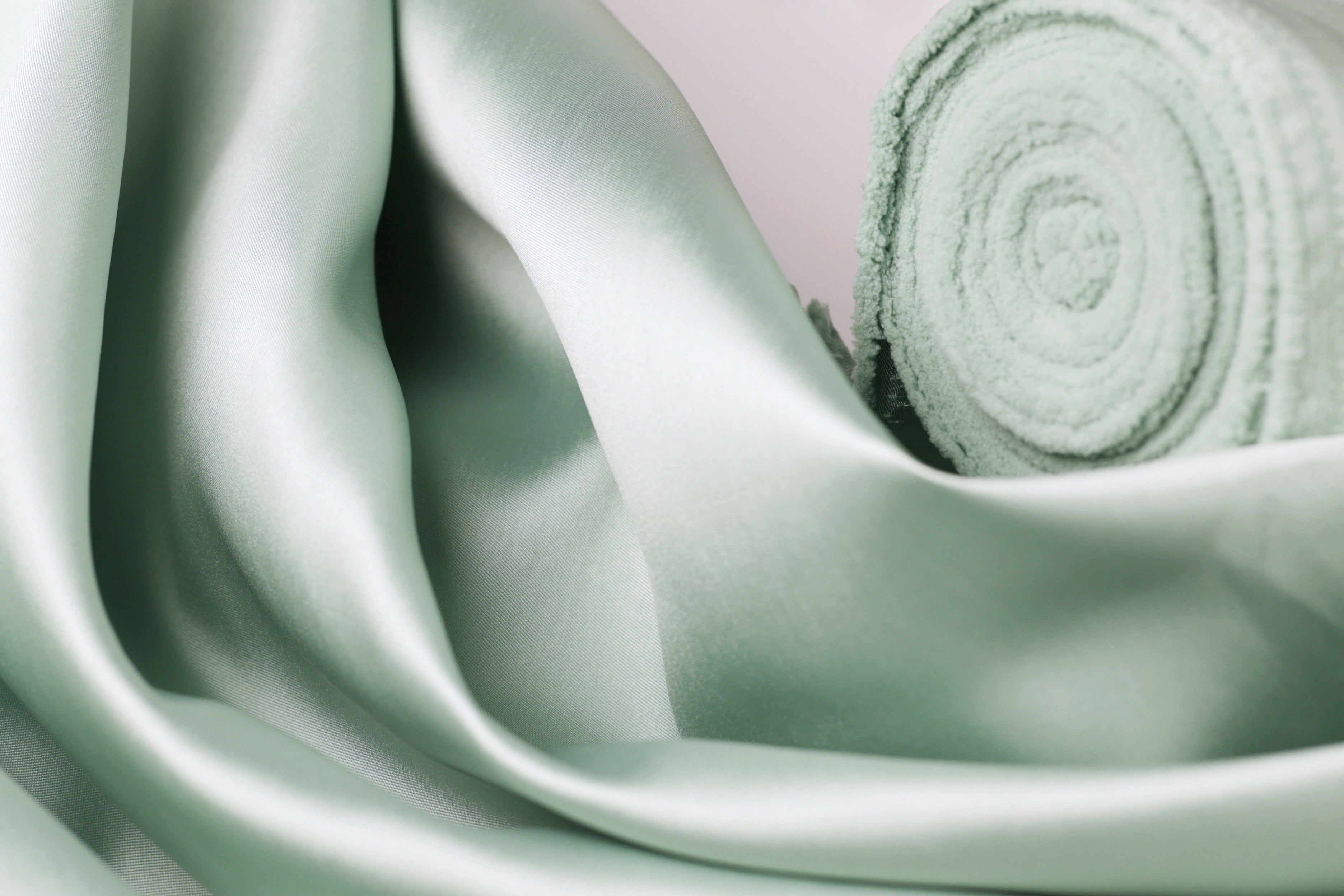 A close-up view of a soft, mint green satin fabric, with a rolled-up piece of the fabric visible in the background.
