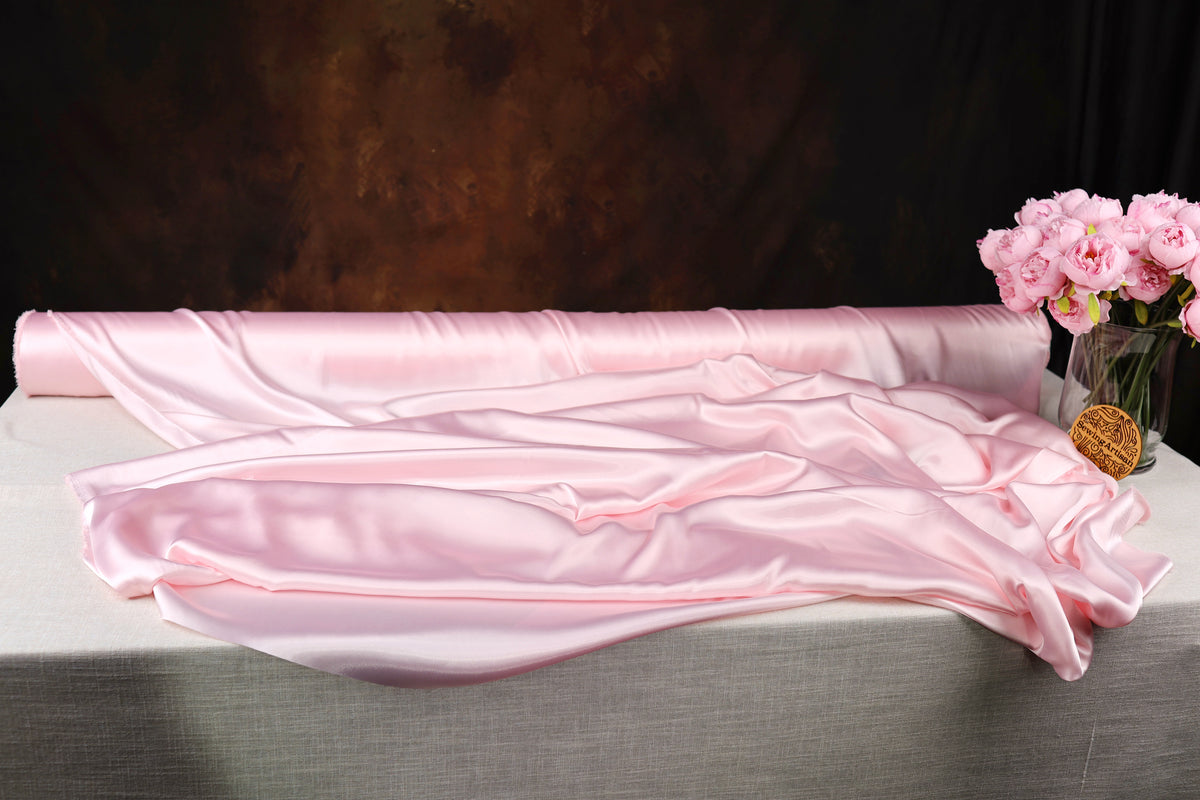a large roll of pink satin fabric, a vase of pink flowers, and a dark background.