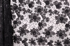 a close-up view of a black and white lace pattern with a floral design. The lace appears to be made of a delicate material, possibly silk or chiffon, and is adorned with numerous small black flowers or leaves. The pattern is intricate and detailed, showcasing the craftsmanship and artistry involved in creating this type of lace.