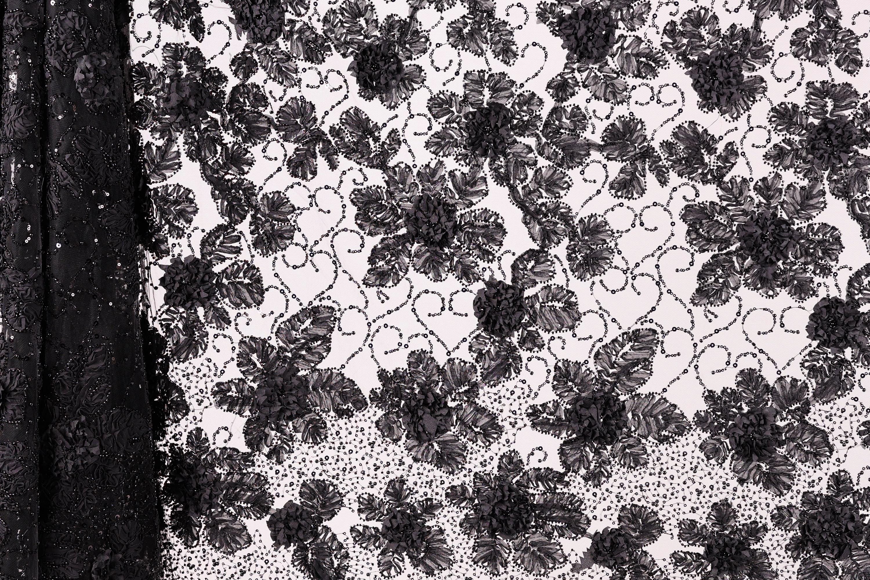a close-up view of a black and white lace pattern with a floral design. The lace appears to be made of a delicate material, possibly silk or chiffon, and is adorned with numerous small black flowers or leaves. The pattern is intricate and detailed, showcasing the craftsmanship and artistry involved in creating this type of lace.