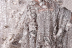 a close-up view of a delicate, intricately designed fabric with a silver sequin pattern. The fabric appears to be a piece of clothing, possibly a dress or a shawl, with a floral or leaf-like design. The background is blurred, drawing focus to the detailed fabric texture and pattern.