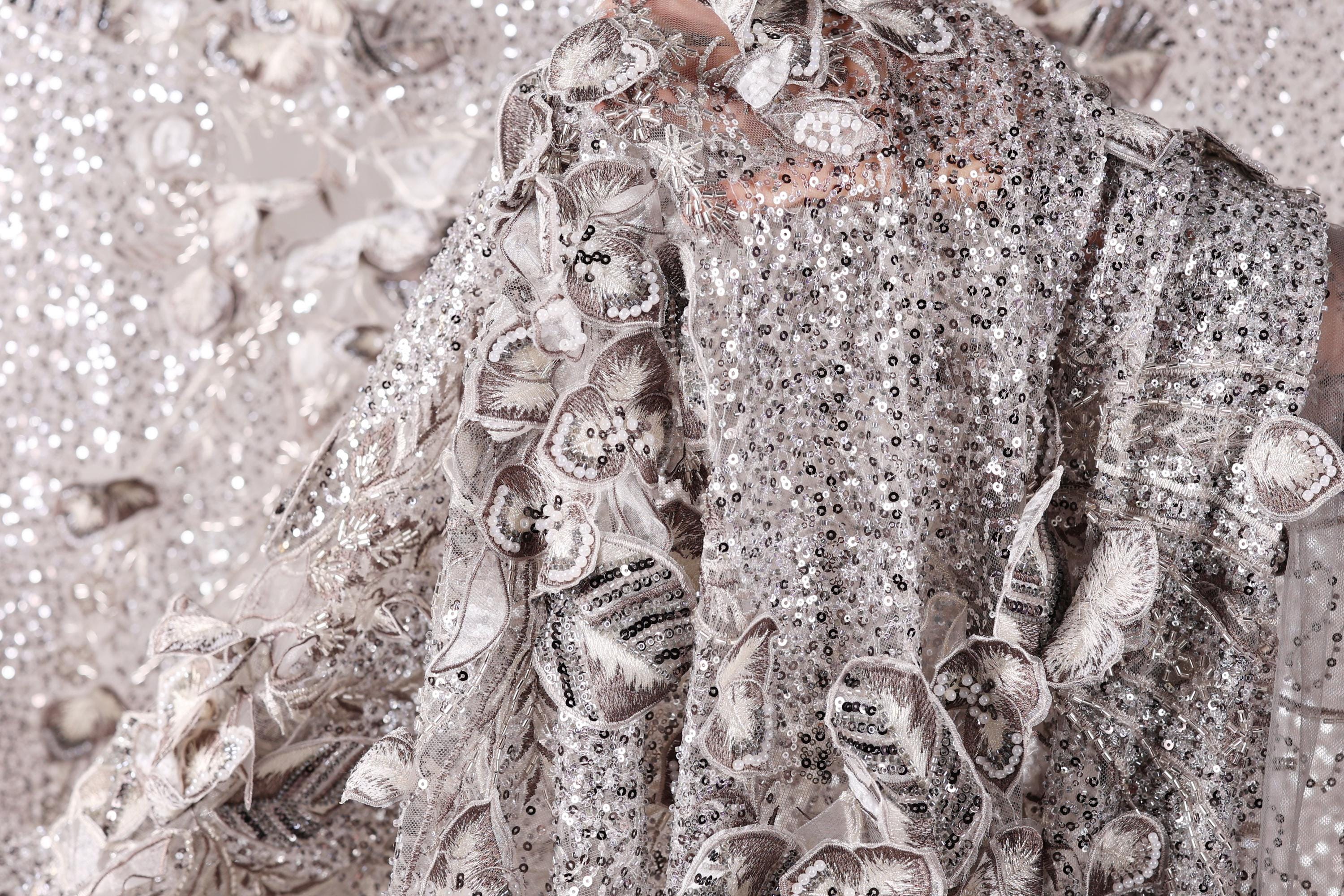a close-up view of a delicate, intricately designed fabric with a silver sequin pattern. The fabric appears to be a piece of clothing, possibly a dress or a shawl, with a floral or leaf-like design. The background is blurred, drawing focus to the detailed fabric texture and pattern.