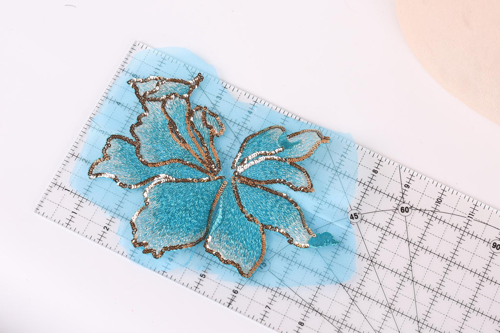 a blue and gold sequin flower design on a transparent surface, with a ruler for scale.