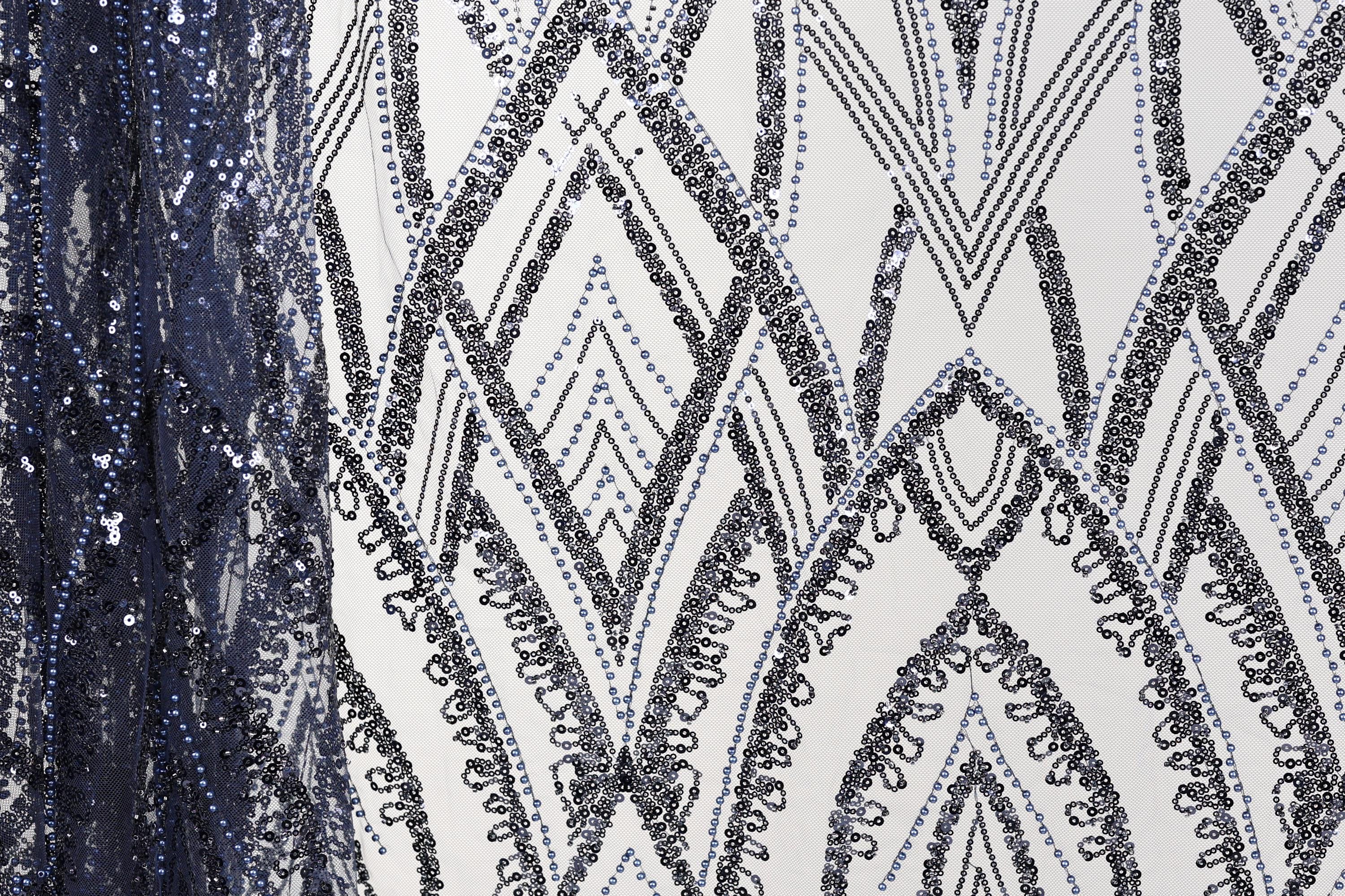 a close-up view of a fabric with a repeating pattern of black and white diamond shapes, interspersed with small blue sequins. The pattern is intricate and visually striking, with the contrasting colors and shapes creating a sense of depth and texture.