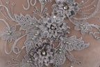 The image is a close-up of a delicate, intricate piece of lace or fabric adorned with silver and white embellishments, including flowers and pearls.