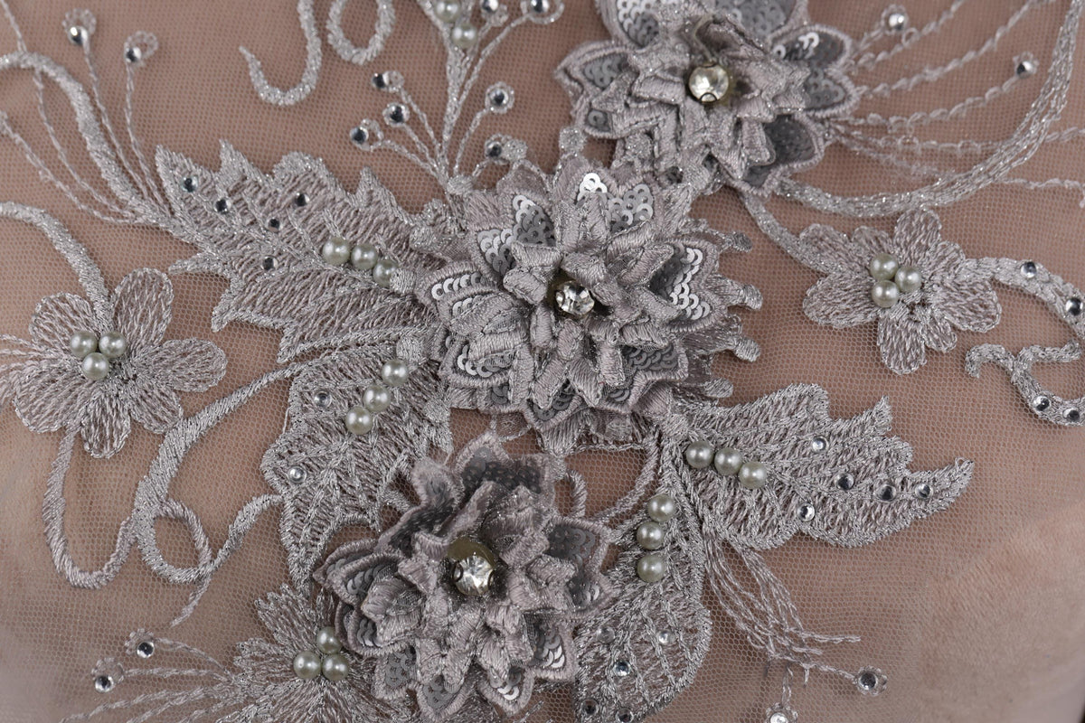 The image is a close-up of a delicate, intricate piece of lace or fabric adorned with silver and white embellishments, including flowers and pearls.