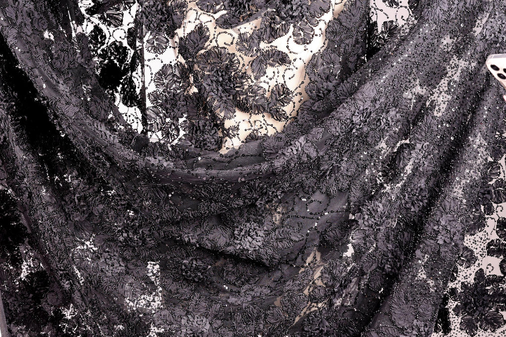 The image is a close-up view of a black and white fabric with a floral pattern. The fabric appears to be made of lace or a similar material, with a mix of black and white colors. The fabric has a textured and intricate appearance, with visible threads or strands running through the fabric.