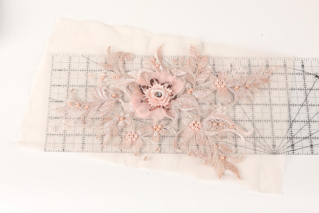 a delicate floral embroidery with a pink flower and leaves, placed on a transparent ruler.