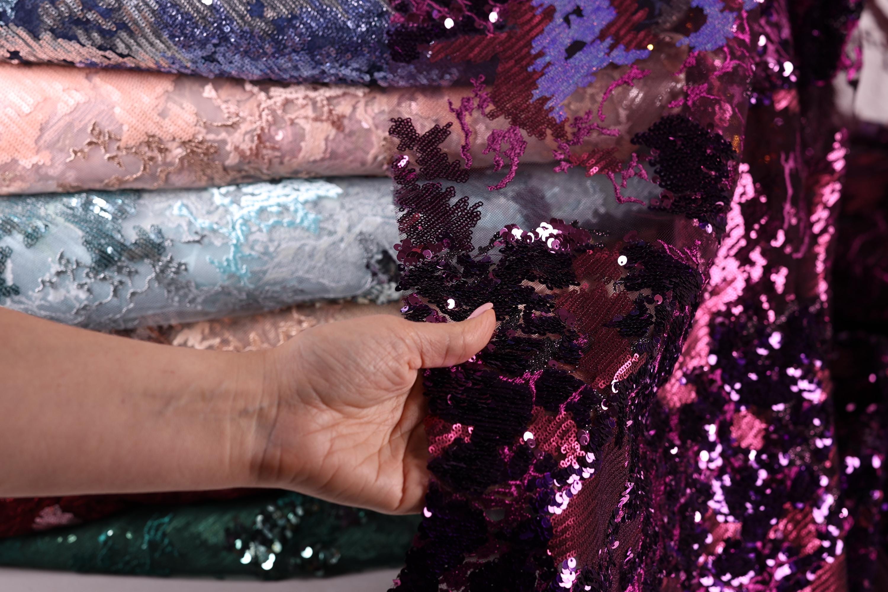 A hand is holding a piece of sequin fabric, which appears to be a vibrant purple color. The hand is positioned in front of a stack of fabric pieces, suggesting that the person is examining or selecting the fabric.