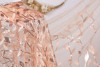 a close-up view of a delicate, peach-colored fabric with a floral pattern. The fabric appears to be made of sheer material, with a light brown color that complements the peach hue. The floral pattern is intricate and detailed, adding a touch of elegance to the fabric. The background is a plain white wall, which serves to highlight the fabric and its design.
