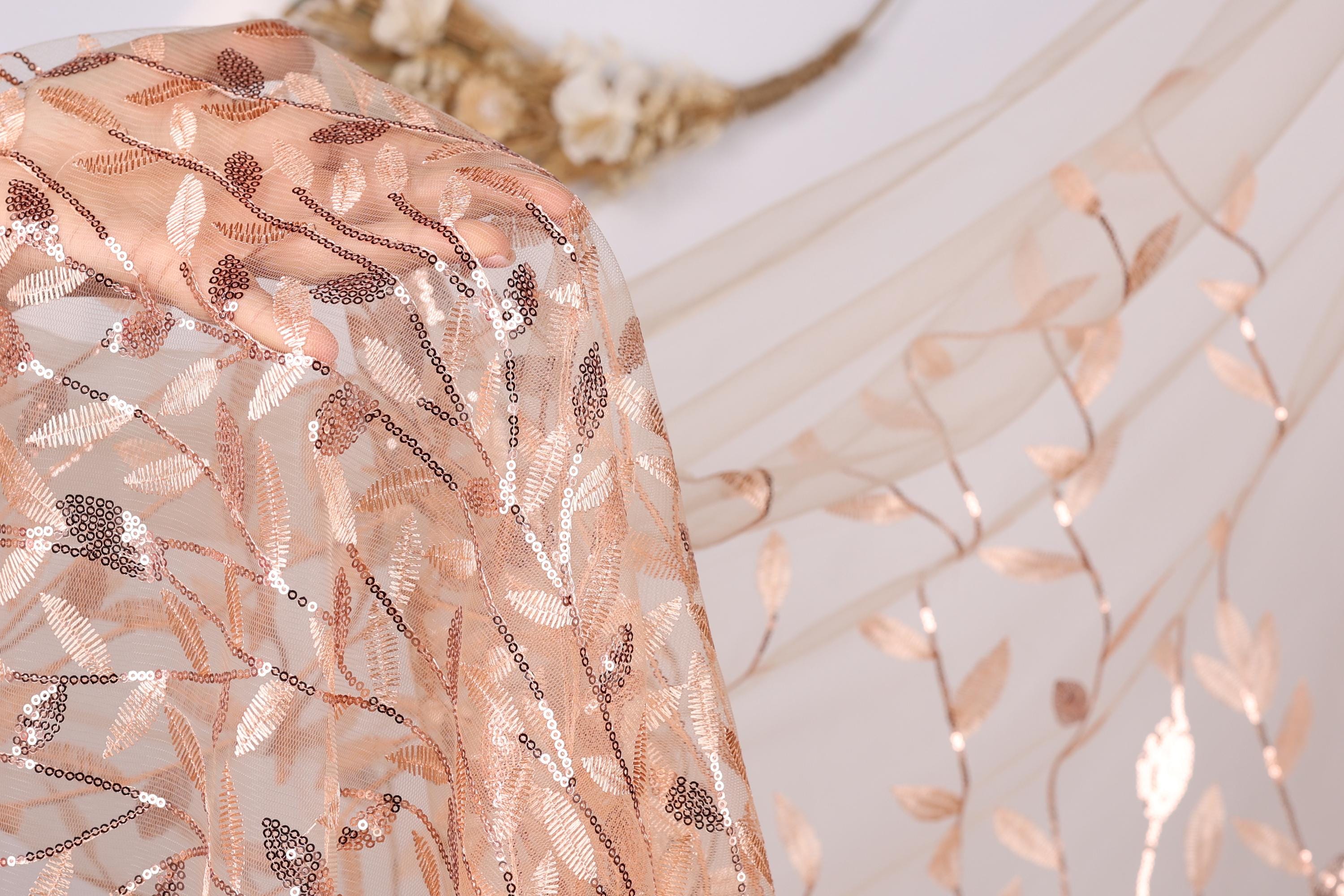 a close-up view of a delicate, peach-colored fabric with a floral pattern. The fabric appears to be made of sheer material, with a light brown color that complements the peach hue. The floral pattern is intricate and detailed, adding a touch of elegance to the fabric. The background is a plain white wall, which serves to highlight the fabric and its design.