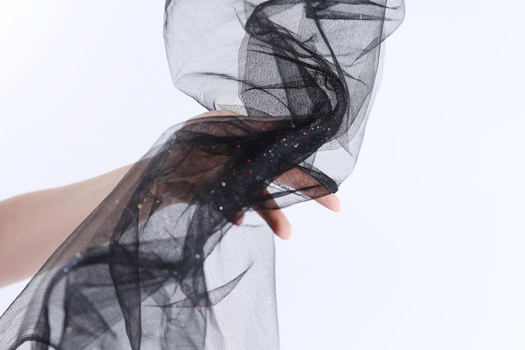 A hand is holding a black, sheer fabric with sparkles on it against a white background.