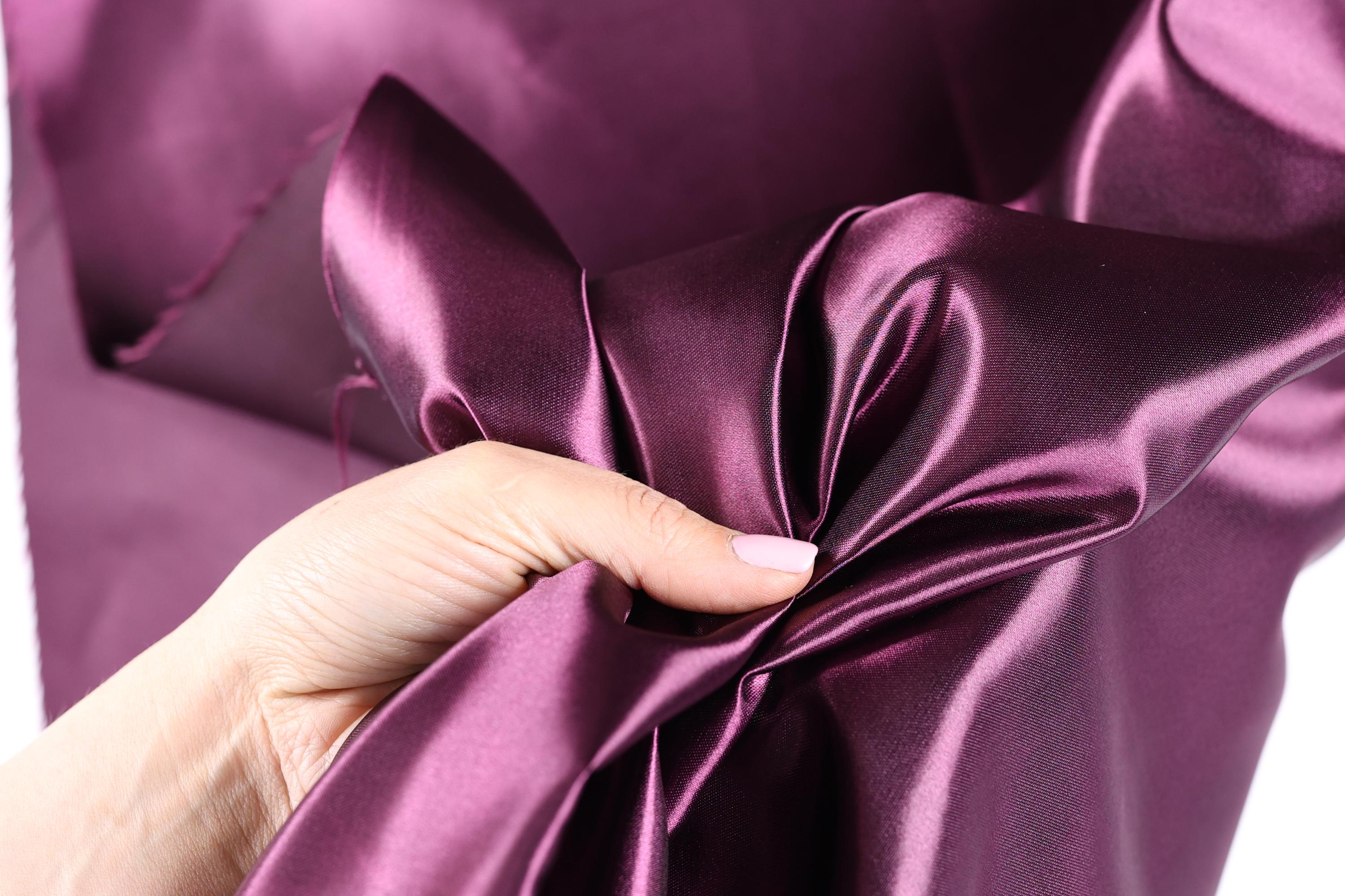A hand is holding a piece of purple satin fabric, examining its texture and appearance.