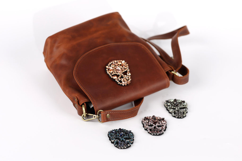 A brown leather handbag with a skull-shaped embellishment, accompanied by four small rhinestone-encrusted charms in various colors.