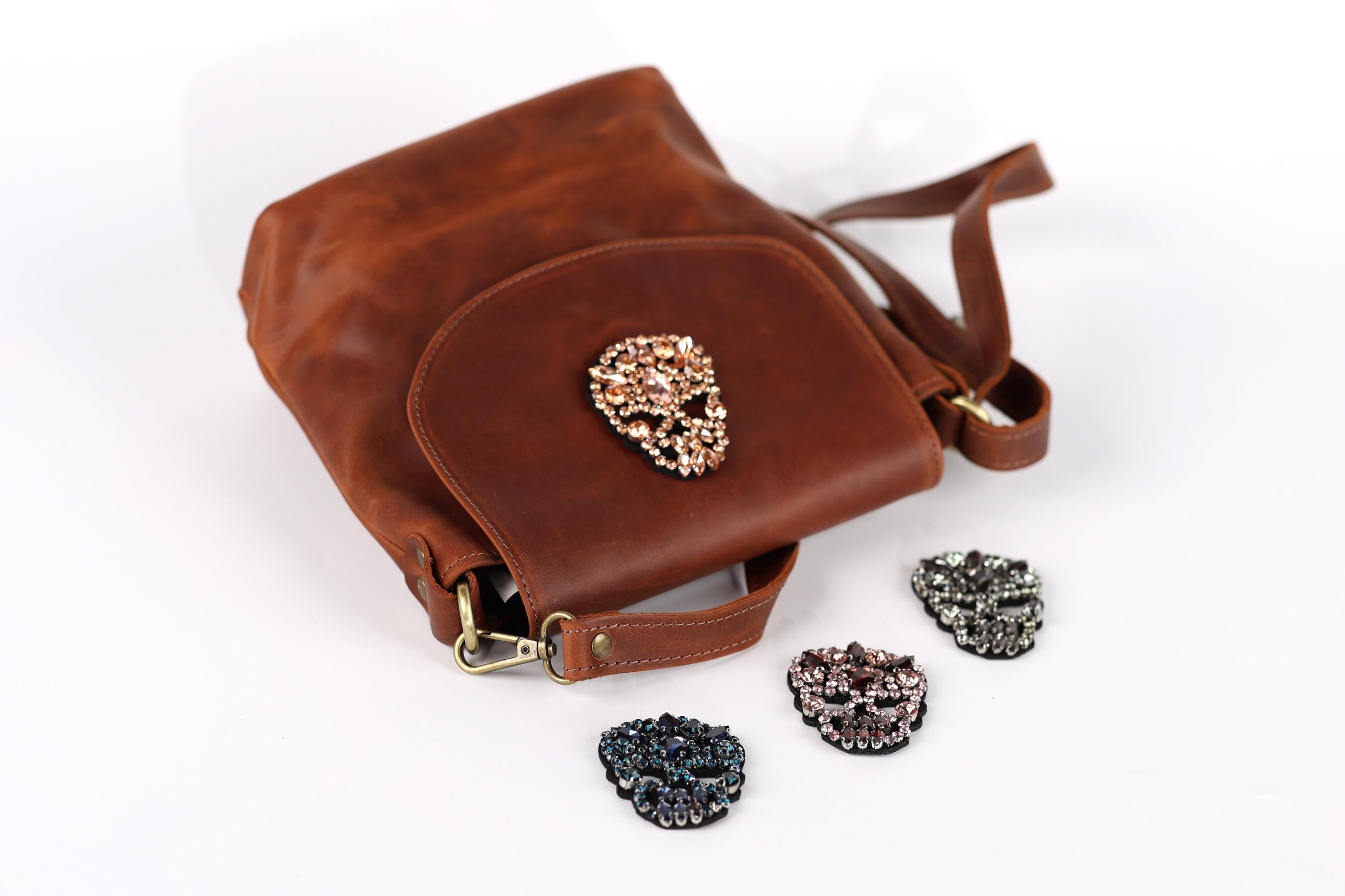 A brown leather handbag with a skull-shaped embellishment, accompanied by four small rhinestone-encrusted charms in various colors.