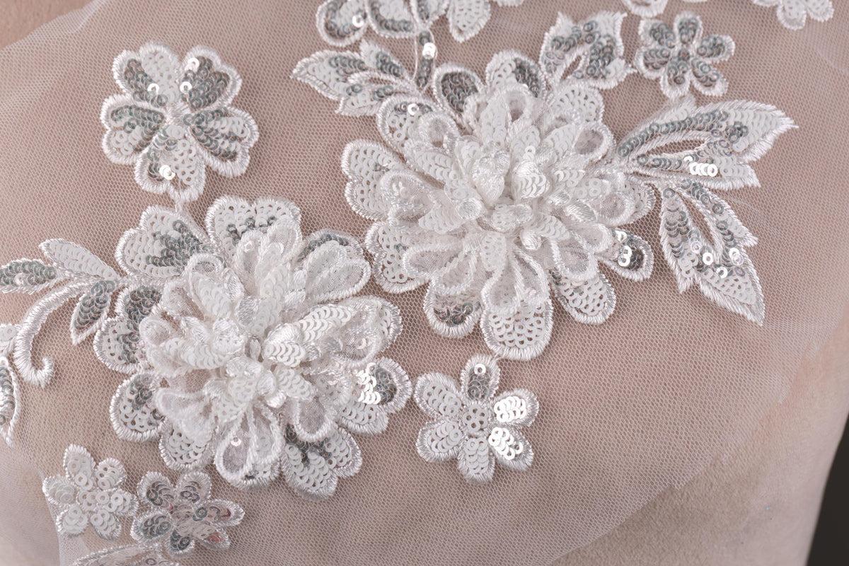 a close-up view of a delicate, intricate white lace design with floral elements. The lace appears to be made of a soft, translucent material, giving it a delicate and elegant appearance. The design features a variety of flowers and leaves, each meticulously crafted and interwoven with the lace, creating a visually stunning and intricate piece of textile art.
