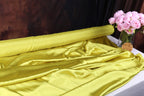a roll of yellow satin fabric on a table, with a vase of pink flowers placed next to it.