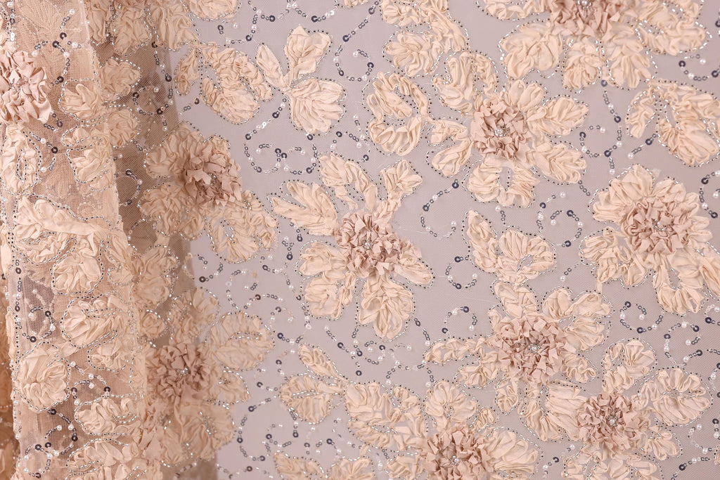 a close-up view of a fabric with a floral pattern, including beige and white flowers and small silver beads. The fabric appears to be made of a delicate, lace-like material, with a subtle shimmer from the silver beads. The image does not contain any text or other discernible objects, and the focus is solely on the intricate floral design.