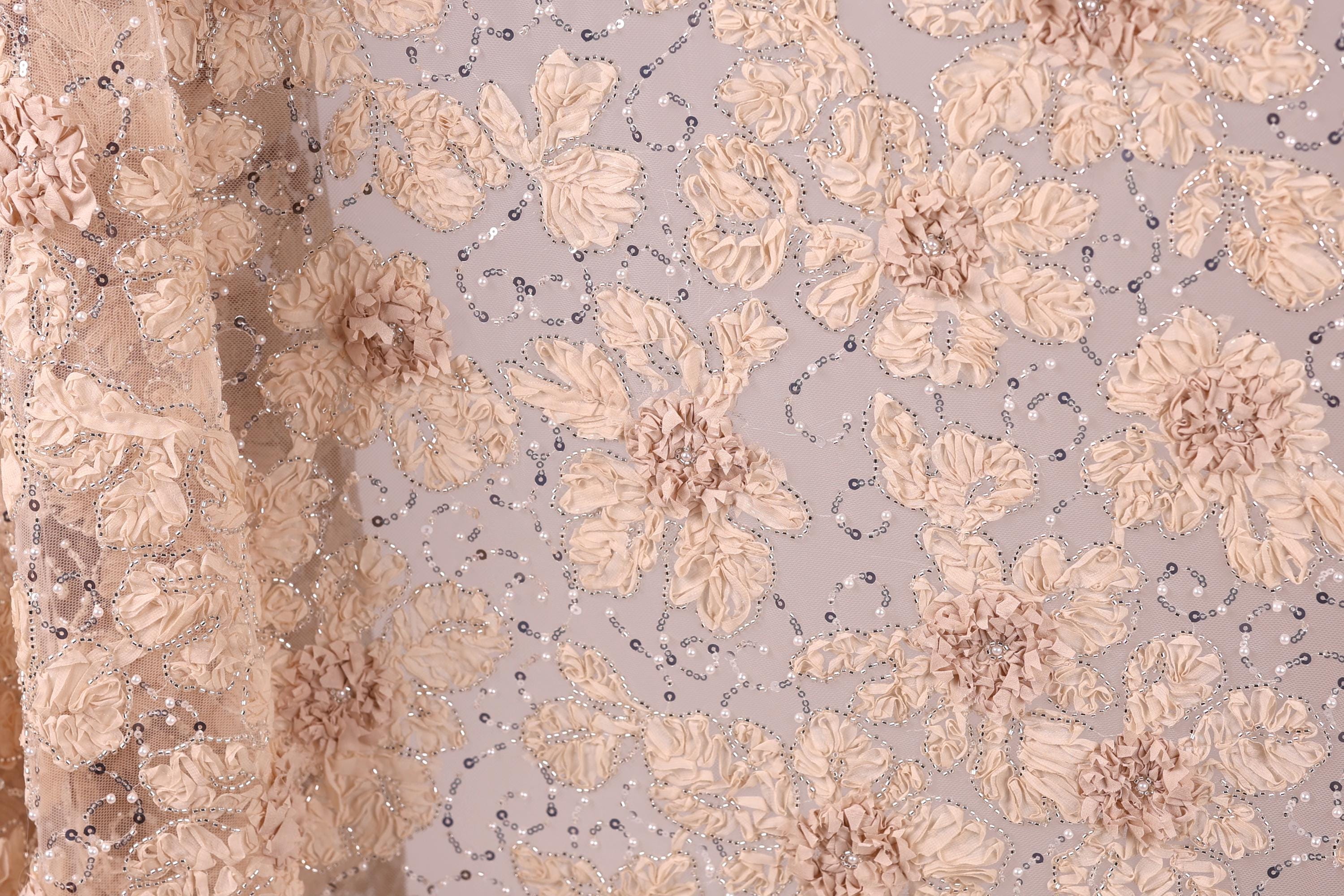 a close-up view of a fabric with a floral pattern, including beige and white flowers and small silver beads. The fabric appears to be made of a delicate, lace-like material, with a subtle shimmer from the silver beads. The image does not contain any text or other discernible objects, and the focus is solely on the intricate floral design.