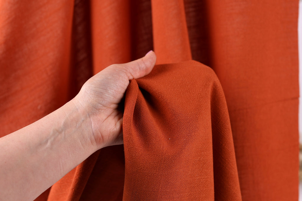 A hand is holding a piece of orange fabric, possibly a curtain or textile, against a red background.