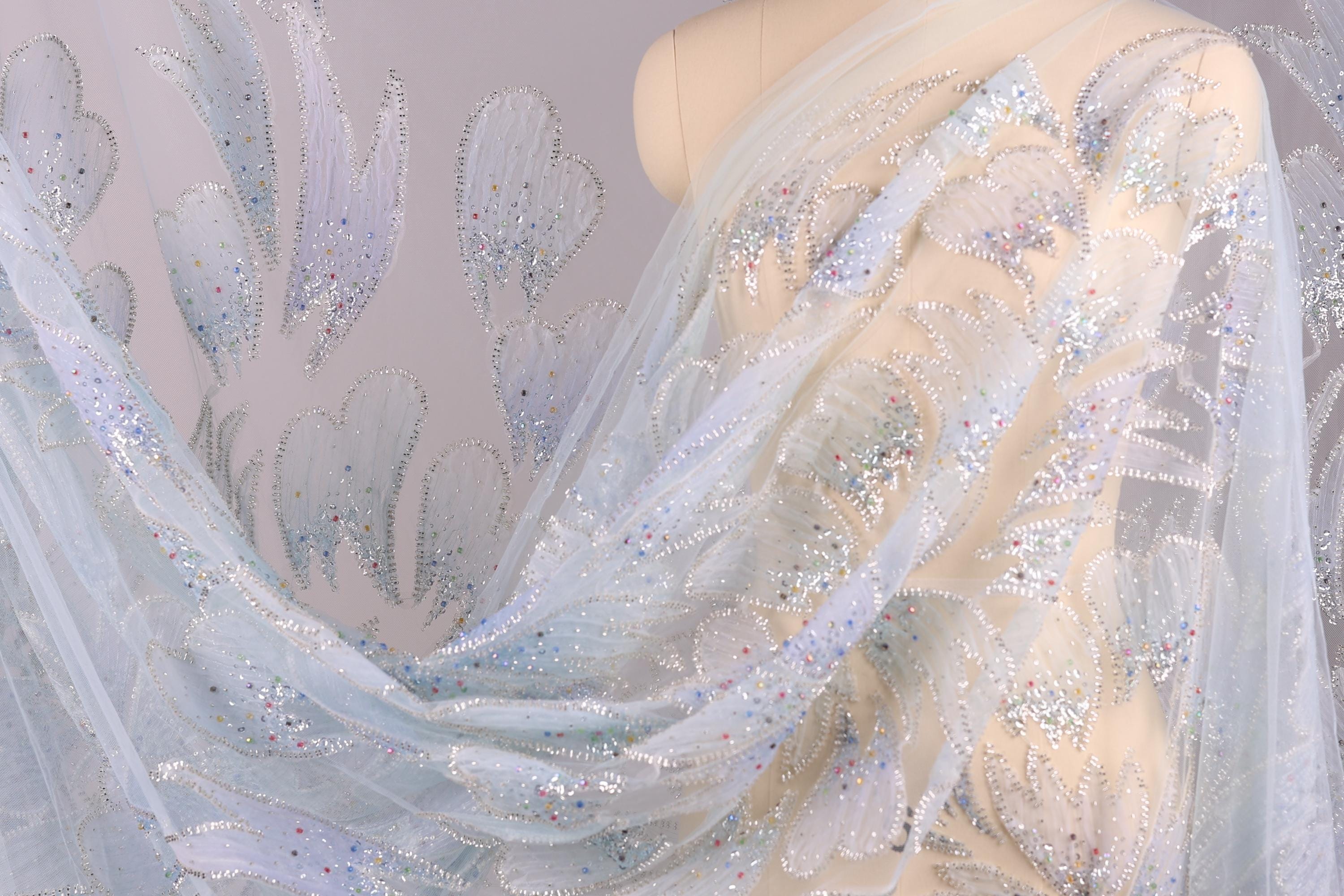 a mannequin wearing a sheer, light blue fabric with a delicate, floral pattern. The fabric appears to be made of sheer material, giving it a delicate and airy appearance. The mannequin is positioned in the center of the image, with the fabric draped over it, showcasing the intricate design and texture of the fabric.