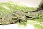 a close-up view of a green, shimmering fabric with a floral pattern. The fabric appears to be made of a sheer material, giving it a delicate and airy appearance. The intricate design and vibrant colors of the fabric create a visually striking contrast against the plain white background.