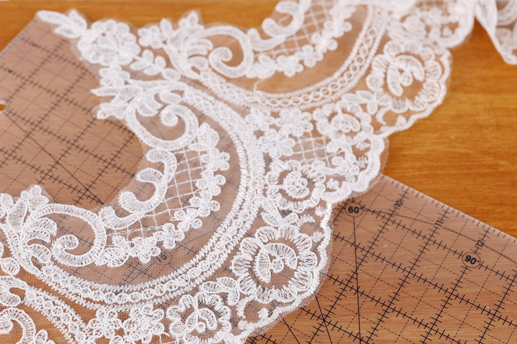 a close-up view of a delicate, intricate white lace fabric with a floral pattern. The lace appears to be handmade and is placed on a wooden surface, possibly a table or a cutting mat. The fabric is spread out, revealing its intricate details and textures.