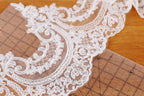 a close-up view of a delicate, intricate white lace fabric with a floral pattern. The lace appears to be handmade and is placed on a wooden surface, possibly a table or a cutting mat. The fabric is spread out, revealing its intricate details and textures.
