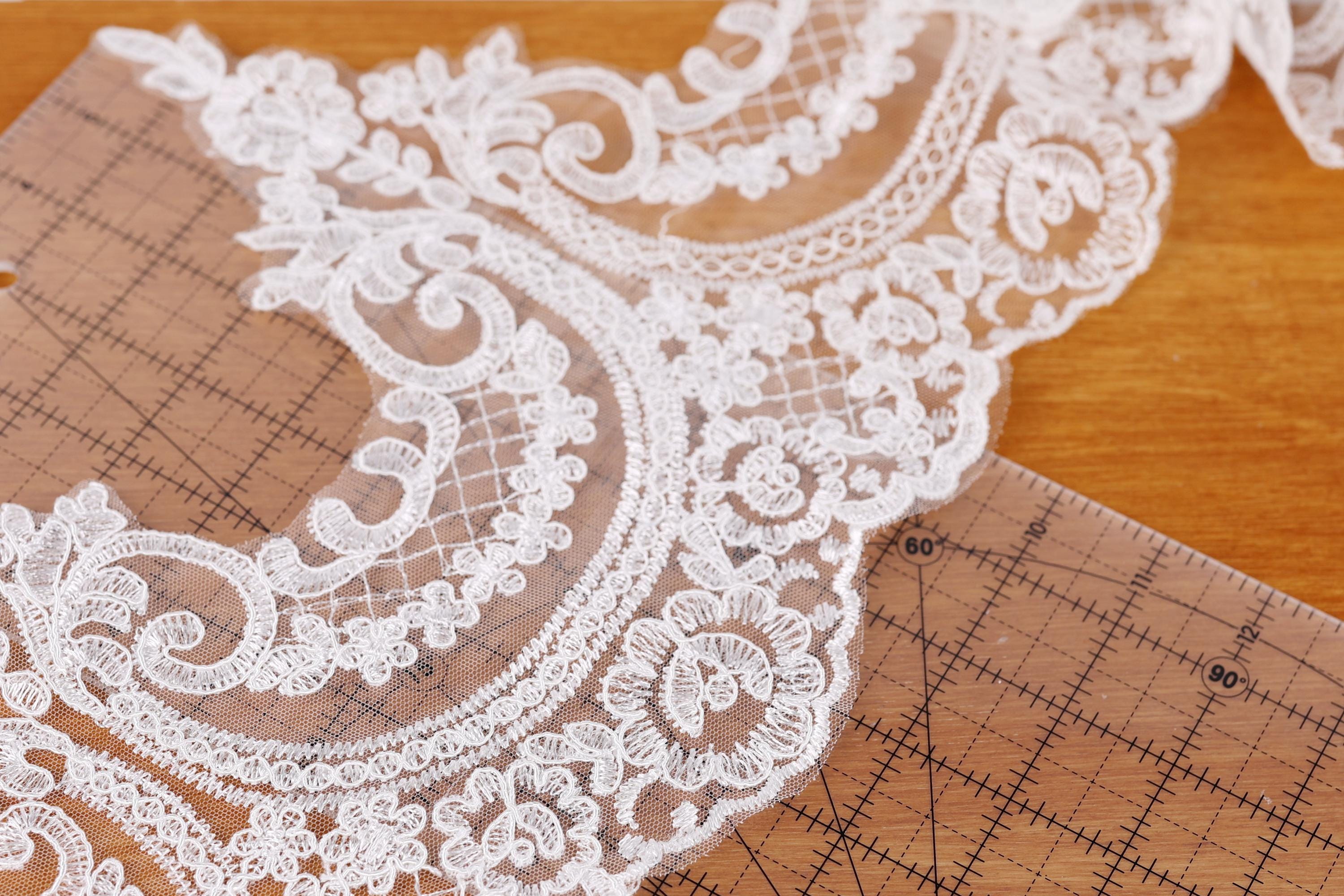 a close-up view of a delicate, intricate white lace fabric with a floral pattern. The lace appears to be handmade and is placed on a wooden surface, possibly a table or a cutting mat. The fabric is spread out, revealing its intricate details and textures.