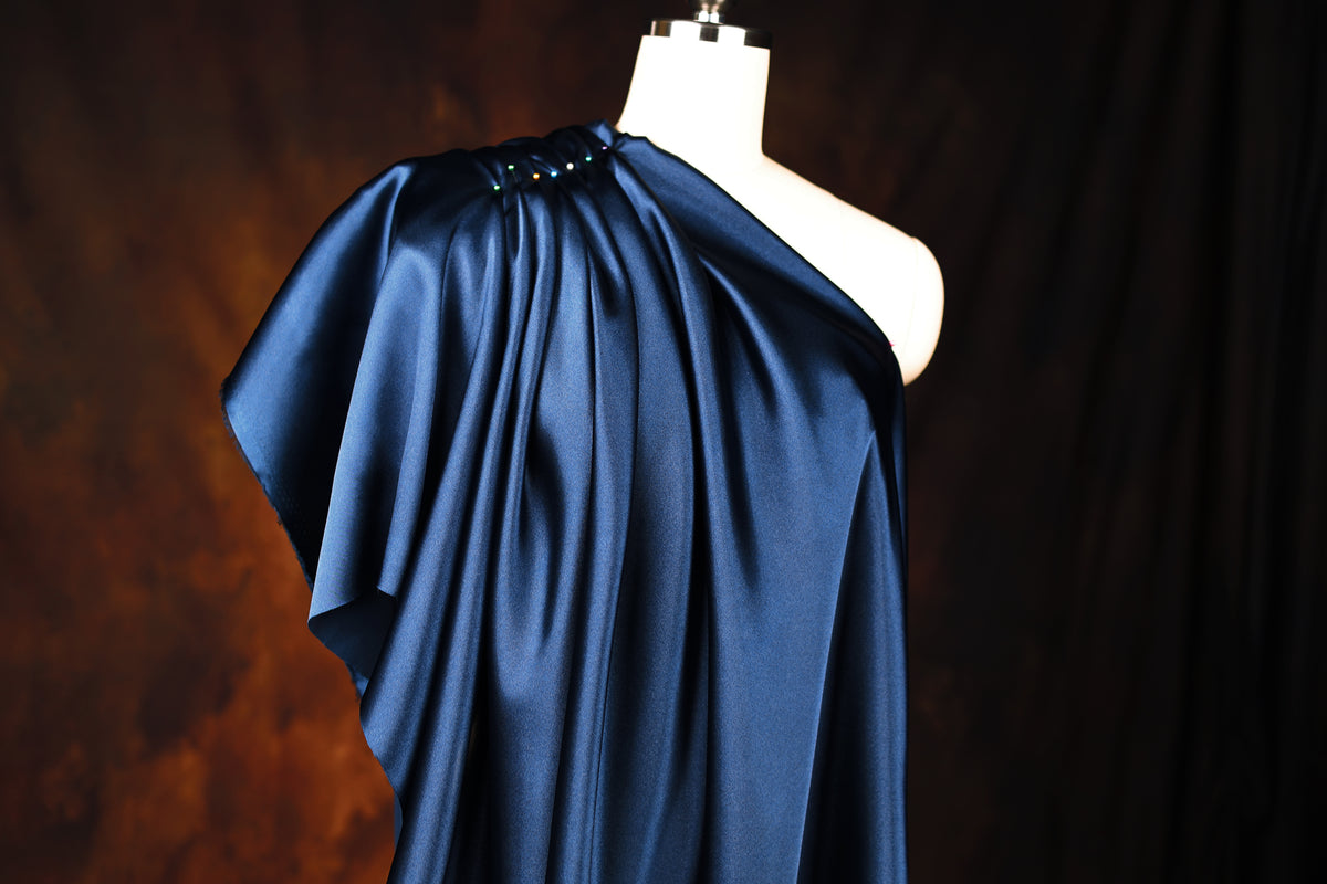 A mannequin wearing a blue satin one-shoulder dress with a draped, flowing silhouette.