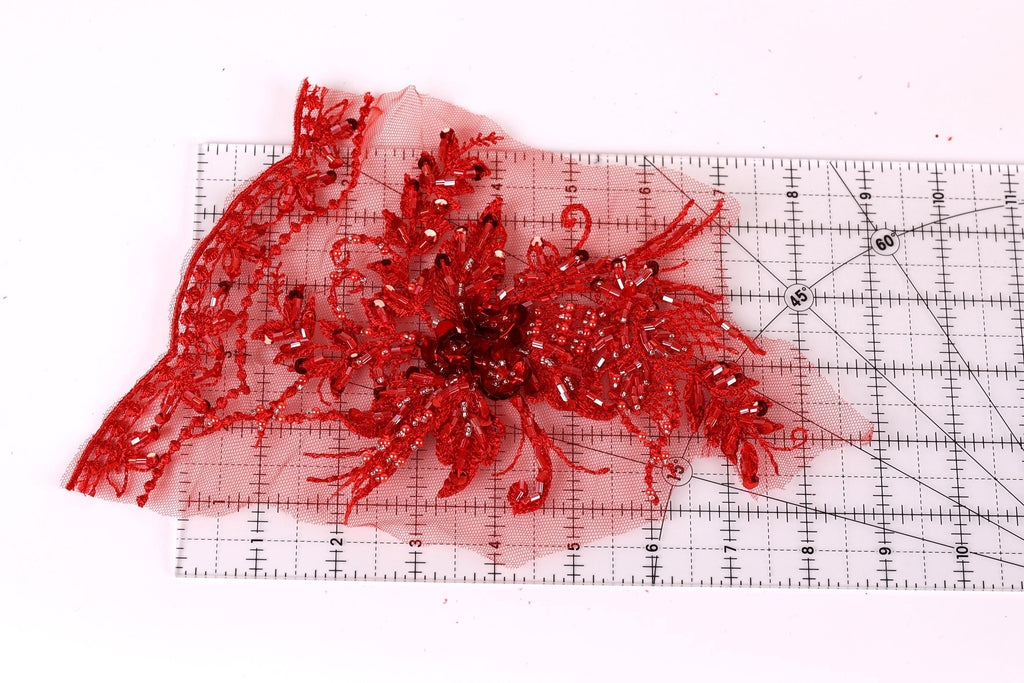 a close-up view of a red, intricately designed fabric with a floral pattern. The fabric appears to be a piece of lace or a similar type of textile, with a vibrant red color and a detailed, decorative design. The fabric is placed on a ruler, which is a common tool used in sewing and crafting to measure and cut fabric accurately.