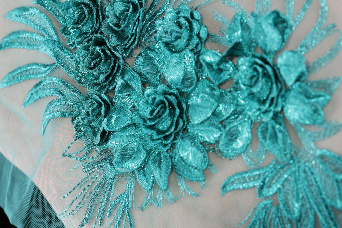 a close-up view of a delicate, intricately embroidered floral design in shades of blue and green. The design is composed of numerous small, shimmering blue flowers and leaves, creating a visually striking and detailed pattern. The flowers and leaves appear to be meticulously crafted, showcasing the skill and artistry of the embroidery work.
