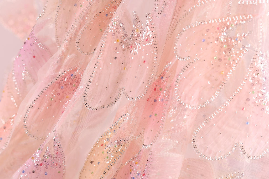 The image is a close-up of a delicate, pink fabric with a sparkling, glittery texture. The fabric appears to be a sheer or sheer-like material, with a floral pattern and small, multicolored beads or sequins scattered throughout the fabric. The image does not contain any text or other discernible objects, and the focus is solely on the intricate details and textures of the fabric.