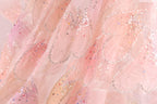 The image is a close-up of a delicate, pink fabric with a sparkling, glittery texture. The fabric appears to be a sheer or sheer-like material, with a floral pattern and small, multicolored beads or sequins scattered throughout the fabric. The image does not contain any text or other discernible objects, and the focus is solely on the intricate details and textures of the fabric.