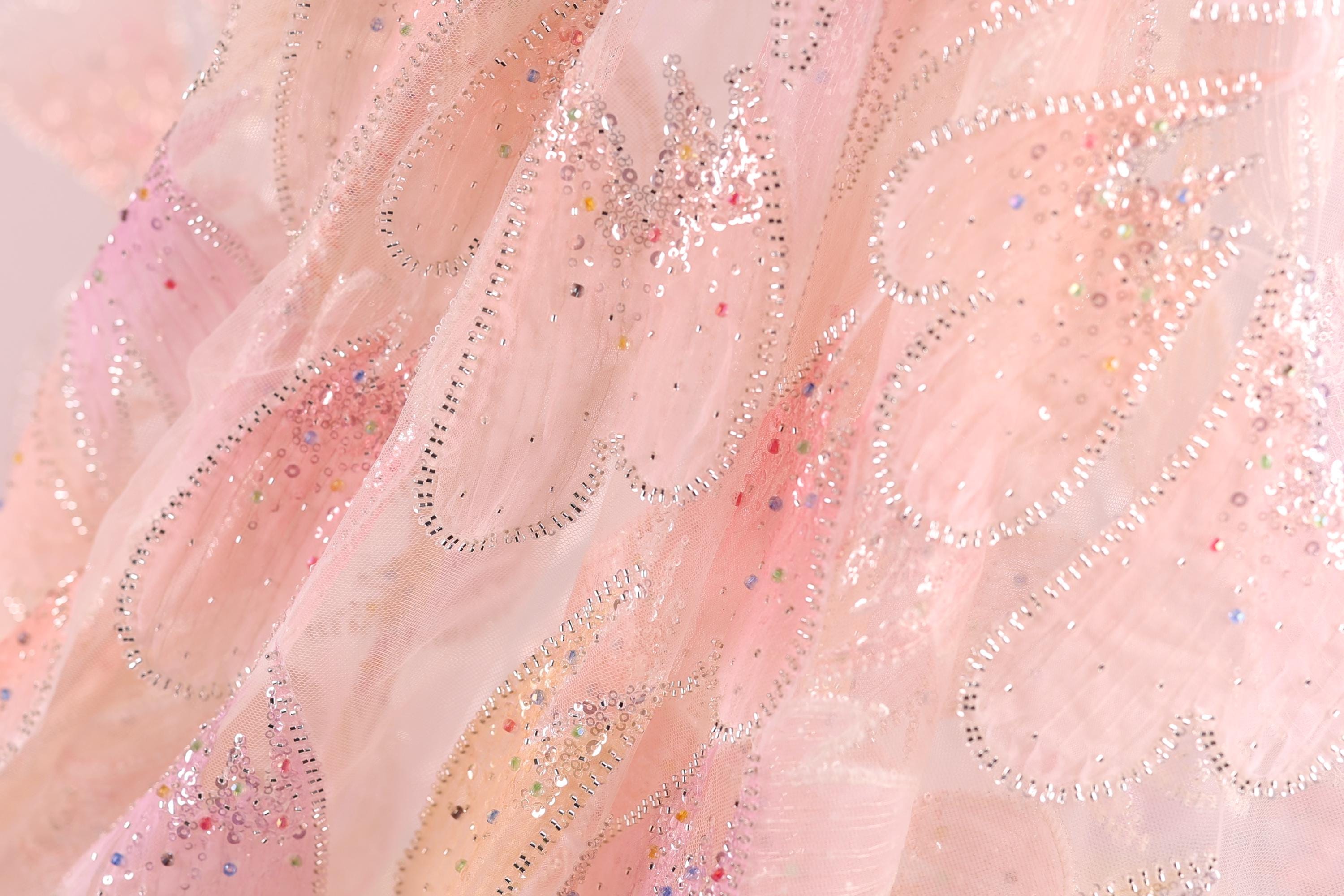 The image is a close-up of a delicate, pink fabric with a sparkling, glittery texture. The fabric appears to be a sheer or sheer-like material, with a floral pattern and small, multicolored beads or sequins scattered throughout the fabric. The image does not contain any text or other discernible objects, and the focus is solely on the intricate details and textures of the fabric.