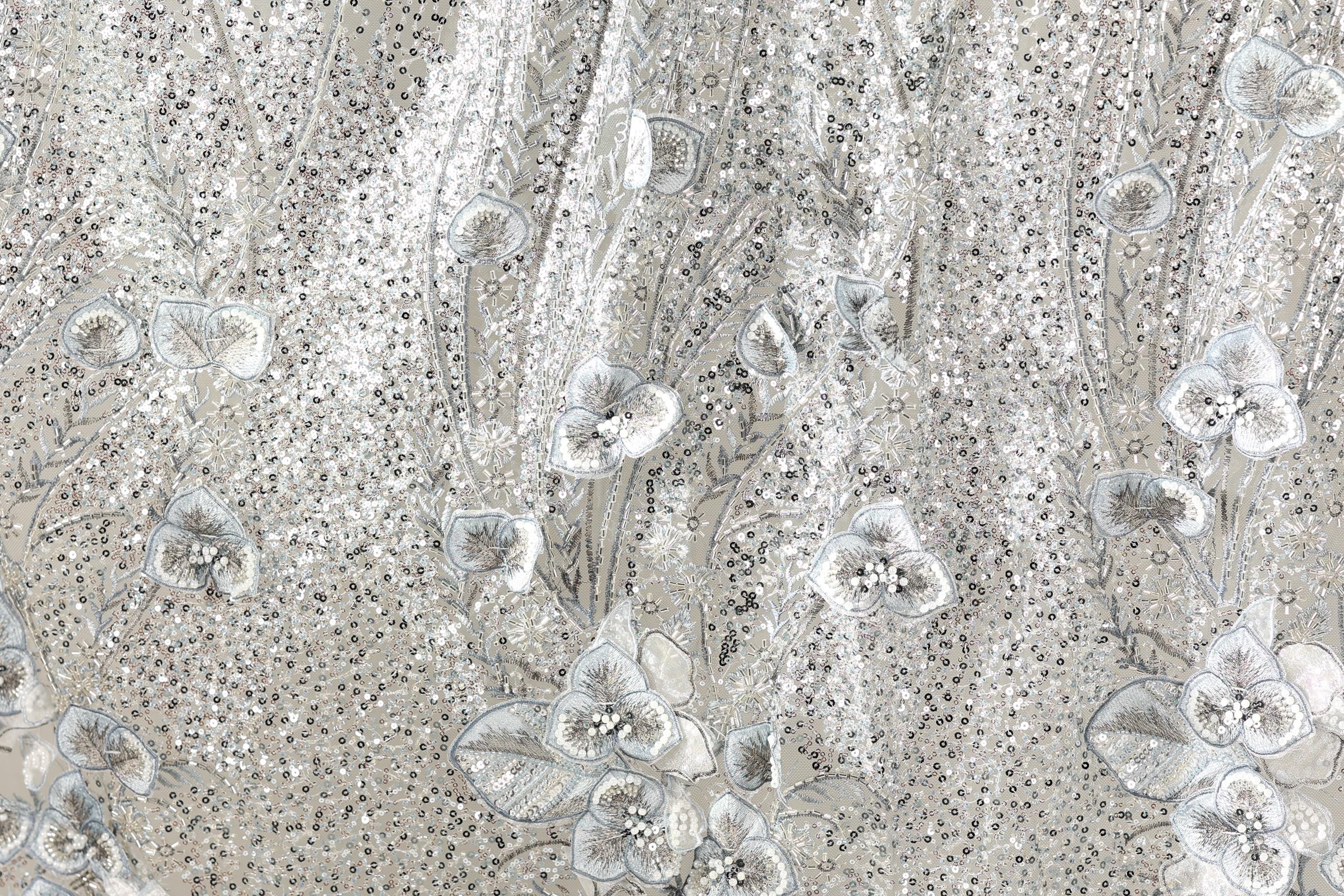 The image is a close-up of a fabric with a floral pattern, featuring small white flowers and leaves. The fabric appears to be made of a shiny, metallic material, giving it a sparkling and elegant appearance.