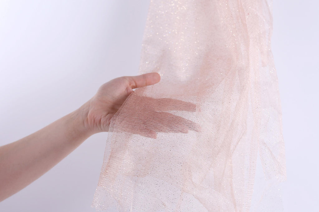 A hand is holding a piece of light pink, sparkly fabric against a white background.