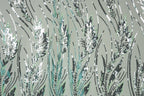 a close-up view of a fabric with a repeating pattern of green and white sequin designs resembling grass or plants. The pattern is intricate and detailed, with the sequins arranged in a way that creates a sense of movement and texture. The overall color scheme is predominantly green and white, with some black and gray accents. The image does not contain any text or other discernible objects, and the focus is solely on the detailed pattern of sequins.