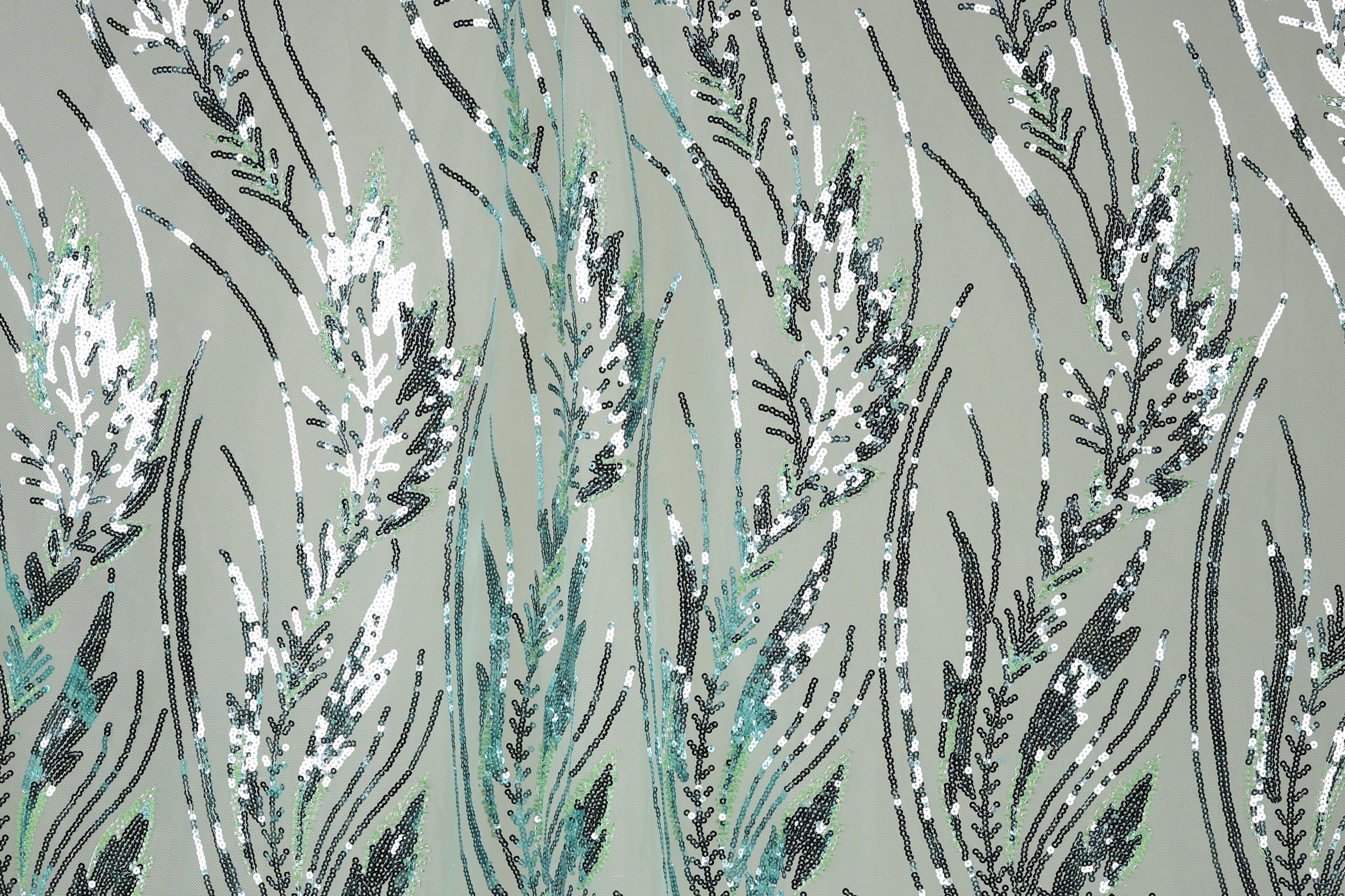 a close-up view of a fabric with a repeating pattern of green and white sequin designs resembling grass or plants. The pattern is intricate and detailed, with the sequins arranged in a way that creates a sense of movement and texture. The overall color scheme is predominantly green and white, with some black and gray accents. The image does not contain any text or other discernible objects, and the focus is solely on the detailed pattern of sequins.