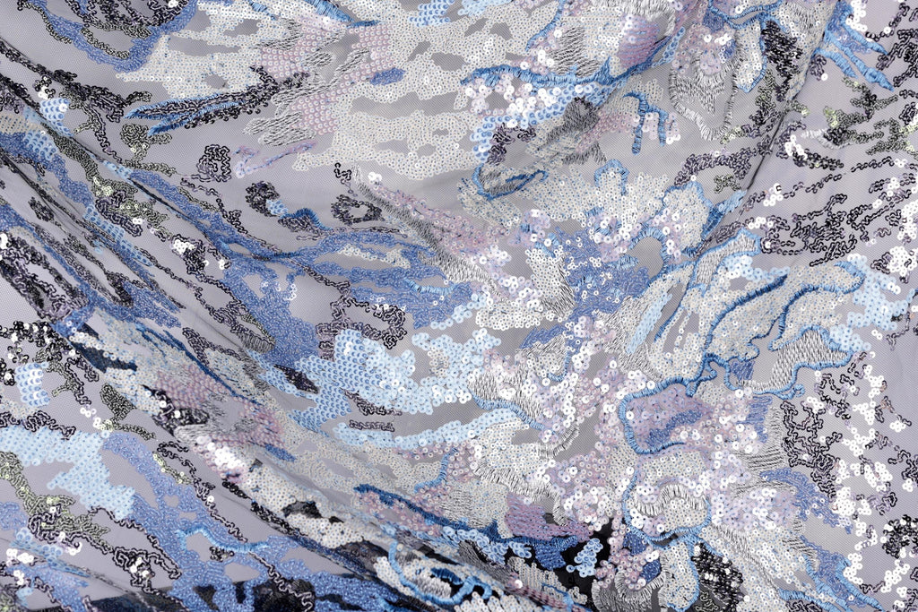 The image is a close-up view of a fabric with a complex, abstract pattern of sequins in various shades of blue, gray, and white. The pattern appears to be a mix of organic and geometric shapes, creating a visually striking and intricate design.
